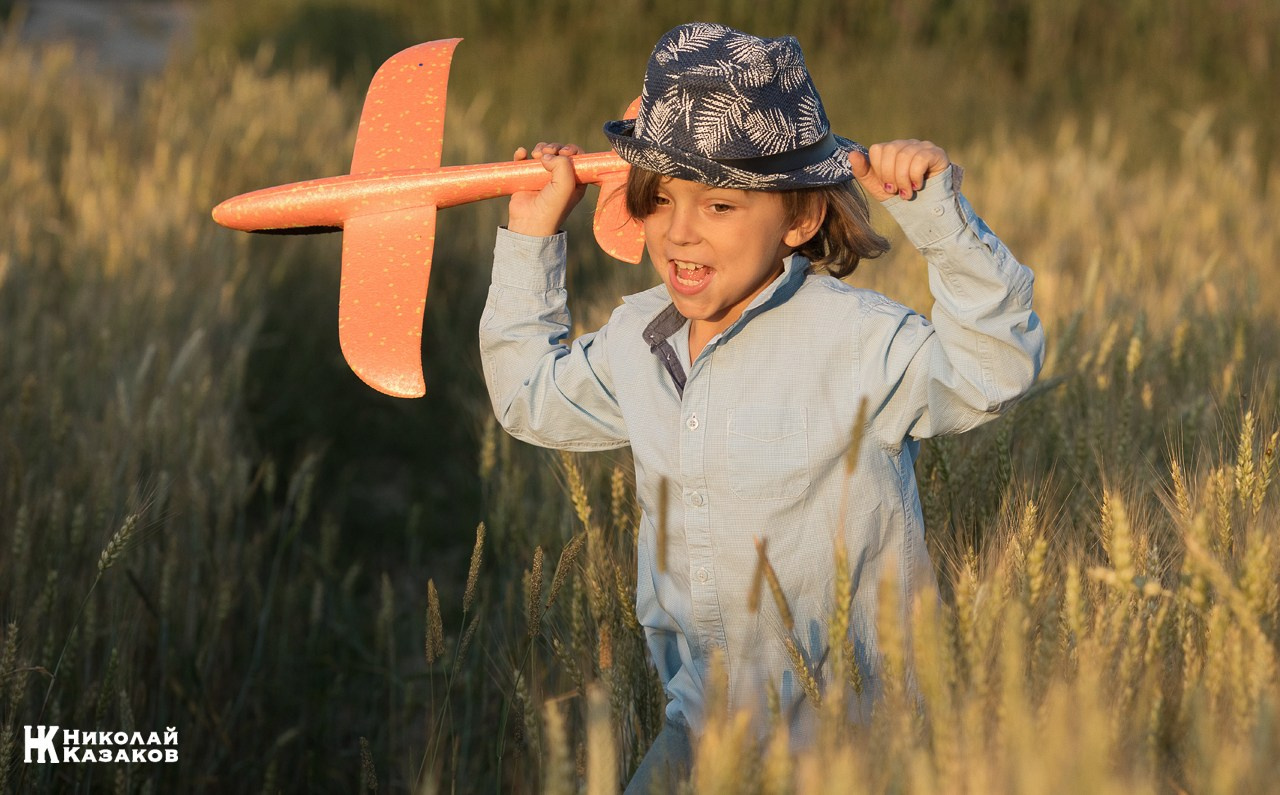 Счастливый маленький мальчик с светлыми волосами бежит по пшеничному полу в лучах закатного солнца , держа в руках игрушечный самолет.