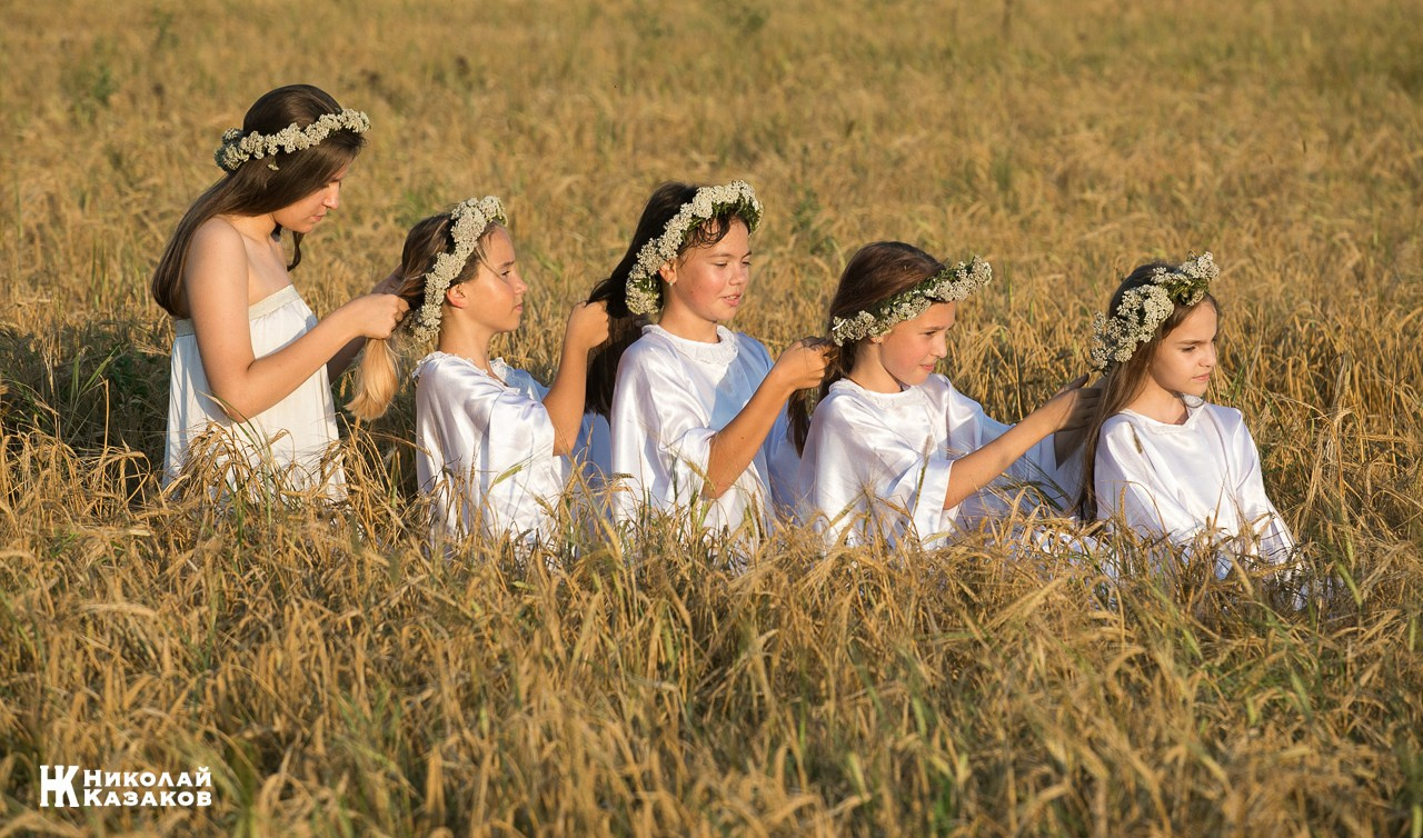 Танцевальная группа девочек подростков  с венками на головах плетут косы сидя в пшенчном поле на закате во время фотосессии