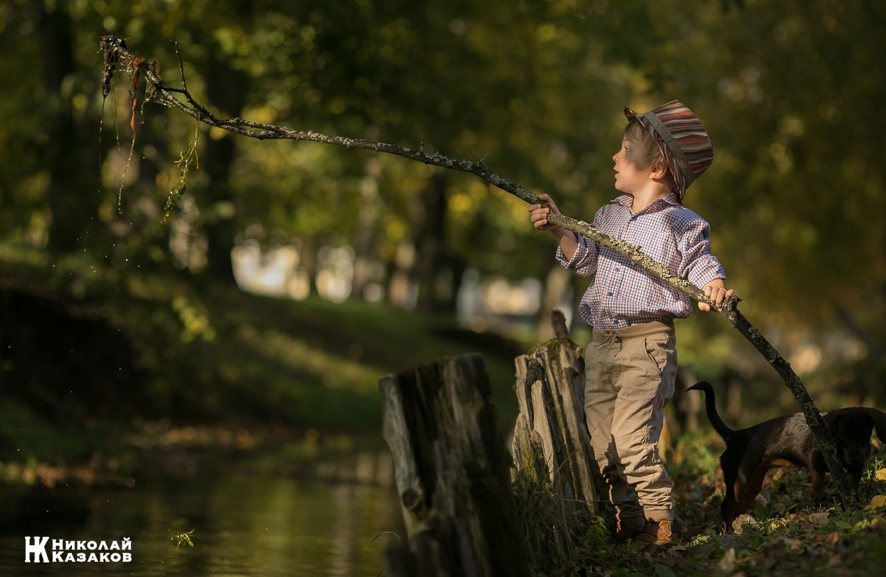 Портрет увлеченного маленького мальчика, ловящего палкой водоросли в речке в лучах утреннего солнца