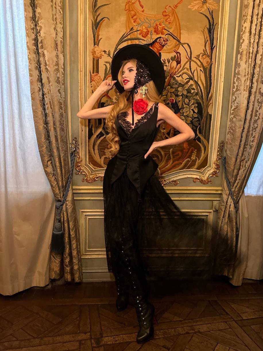 Девушка в черной шляпе и черном платье с кружевным лифом позирует на фоне золотого интерьера