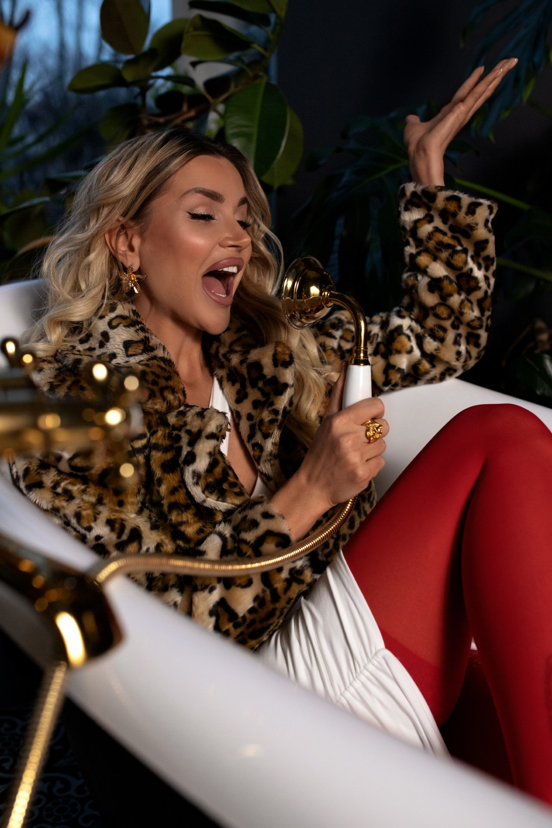 Стильная блондинка в леопардовой шубе и красных колготках сидит в винтажной ванне и поёт в золотую душевую лейку, позируя в роскошном интерьере с растениями в студии