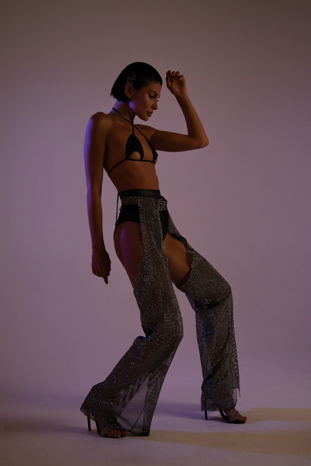 Модель в черном бикини и блестящих прозрачных брюках с высокой талией позирует в студии на фоне мягкой неоновой подсветки.