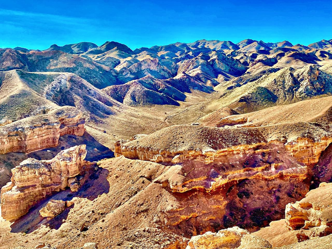Захватывающий вид на Лунный каньон в районе Алматы в Казахстане под ярким синим небом. Пейзаж состоит из красновато-коричневых скал, возвышающихся над долиной, с уникальной текстурой и формами. На заднем плане простираются холмы и горы, создавая глубину и величественность композиции. Цвета насыщенные, с акцентом на контраст между тёплыми тонами земли и холодным небесным оттенком.