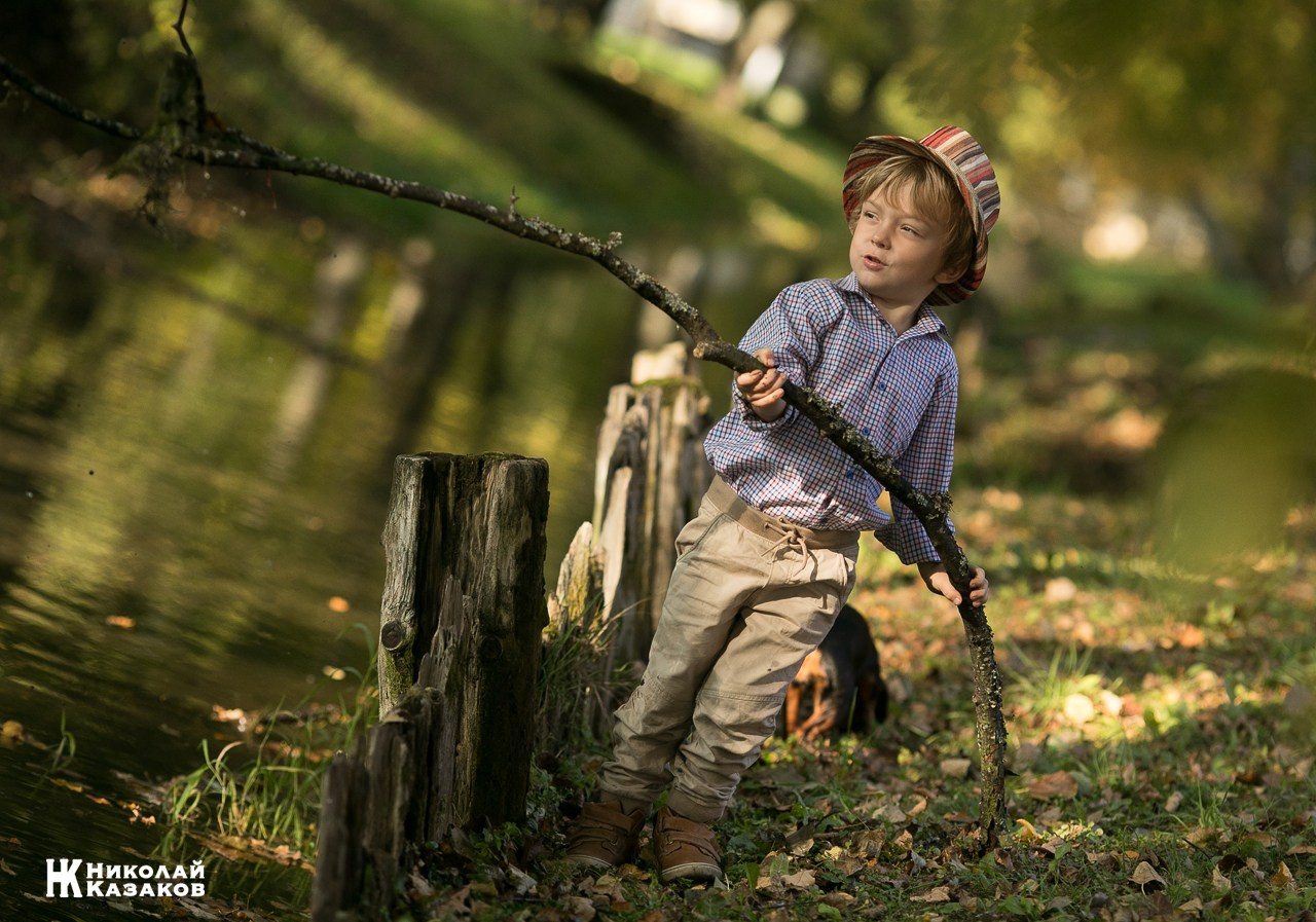 Портрет увлеченного маленького мальчика, ловящего палкой водоросли в речке в лучах утреннего солнца
