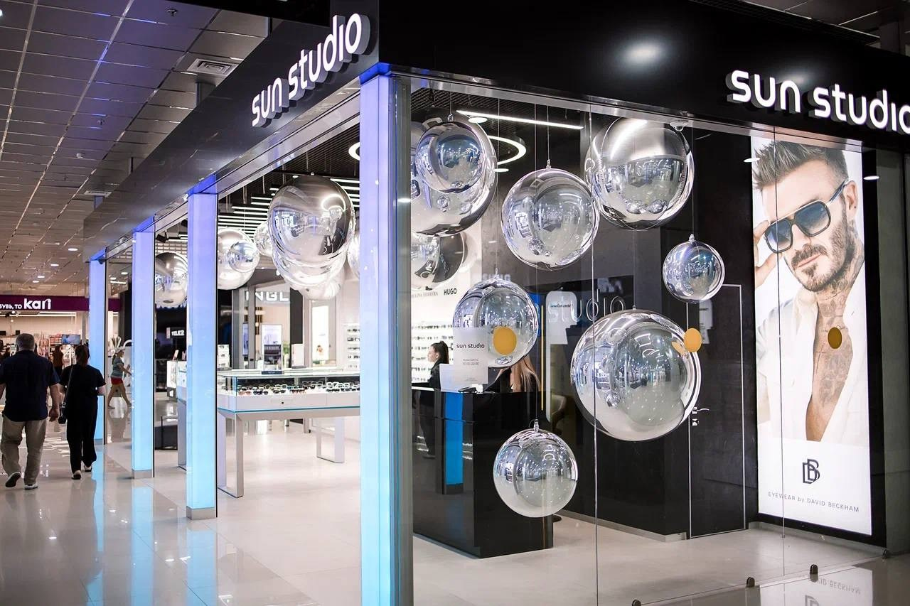 Интерьер современного магазина оптики «Sun Studio» в торговом центре. Входная группа украшена гроздьями крупных зеркальных и прозрачных шаров, подвешенных на разной высоте. Внутри видны подсвеченные стеклянные витрины с очками, стойка рецепции и рекламный баннер с мужским портретом бренда Eyewear by David Beckham. На переднем плане мимо магазина проходят посетители.