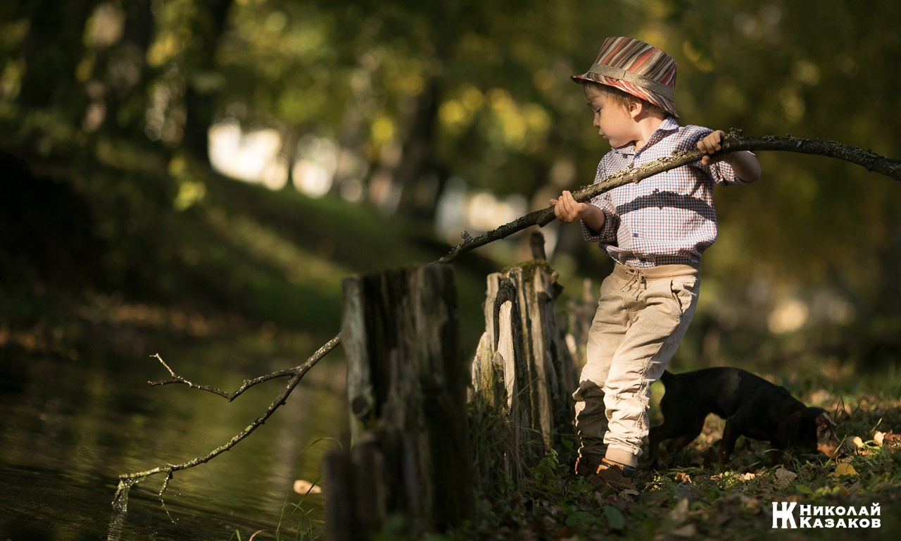 Портрет увлеченного маленького мальчика, ловящего палкой водоросли в речке в лучах утреннего солнца