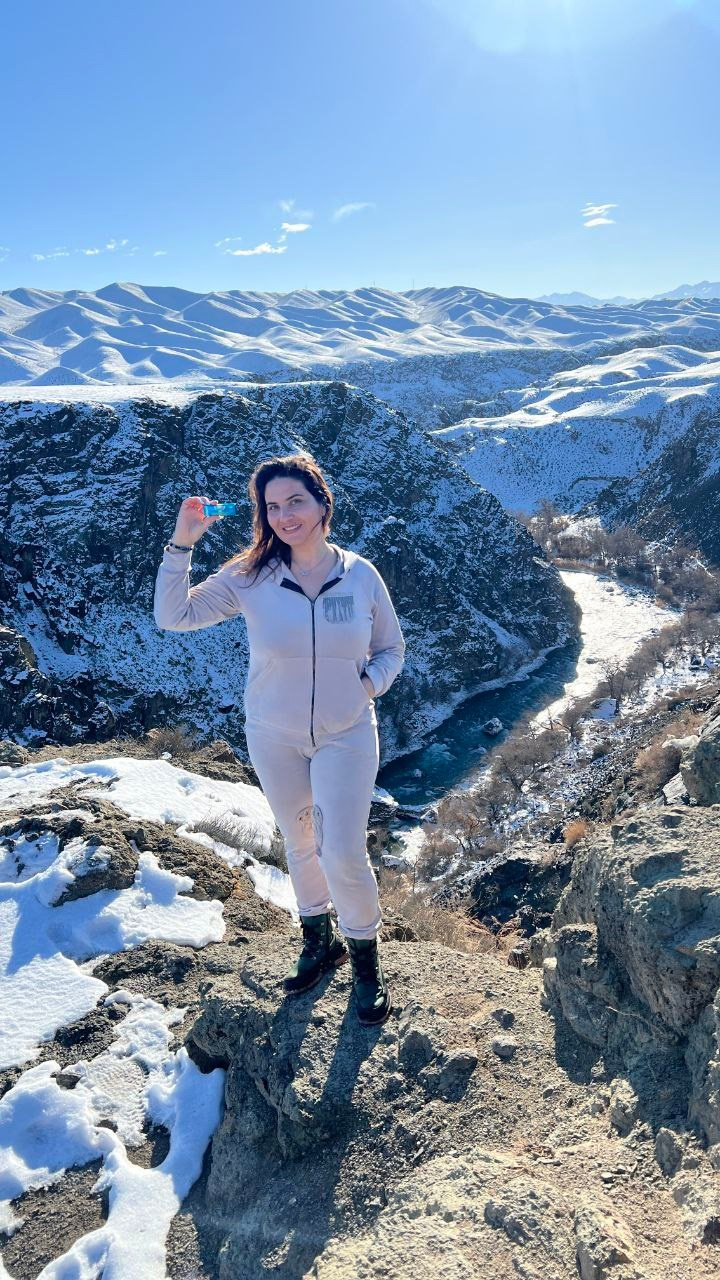 Гид по Алматы Кристина из команды Melon Travel в светлой одежде и ботинках стоит на краю скалистого обрыва. На заднем плане видны заснеженные холмы и горы, а также глубокий каньон с рекой бирюзового цвета. Яркое солнце освещает зимний пейзаж, создавая впечатление холодного, но ясного дня. Женщина держит в руке маленький предмет и улыбается, позируя для фотографии.