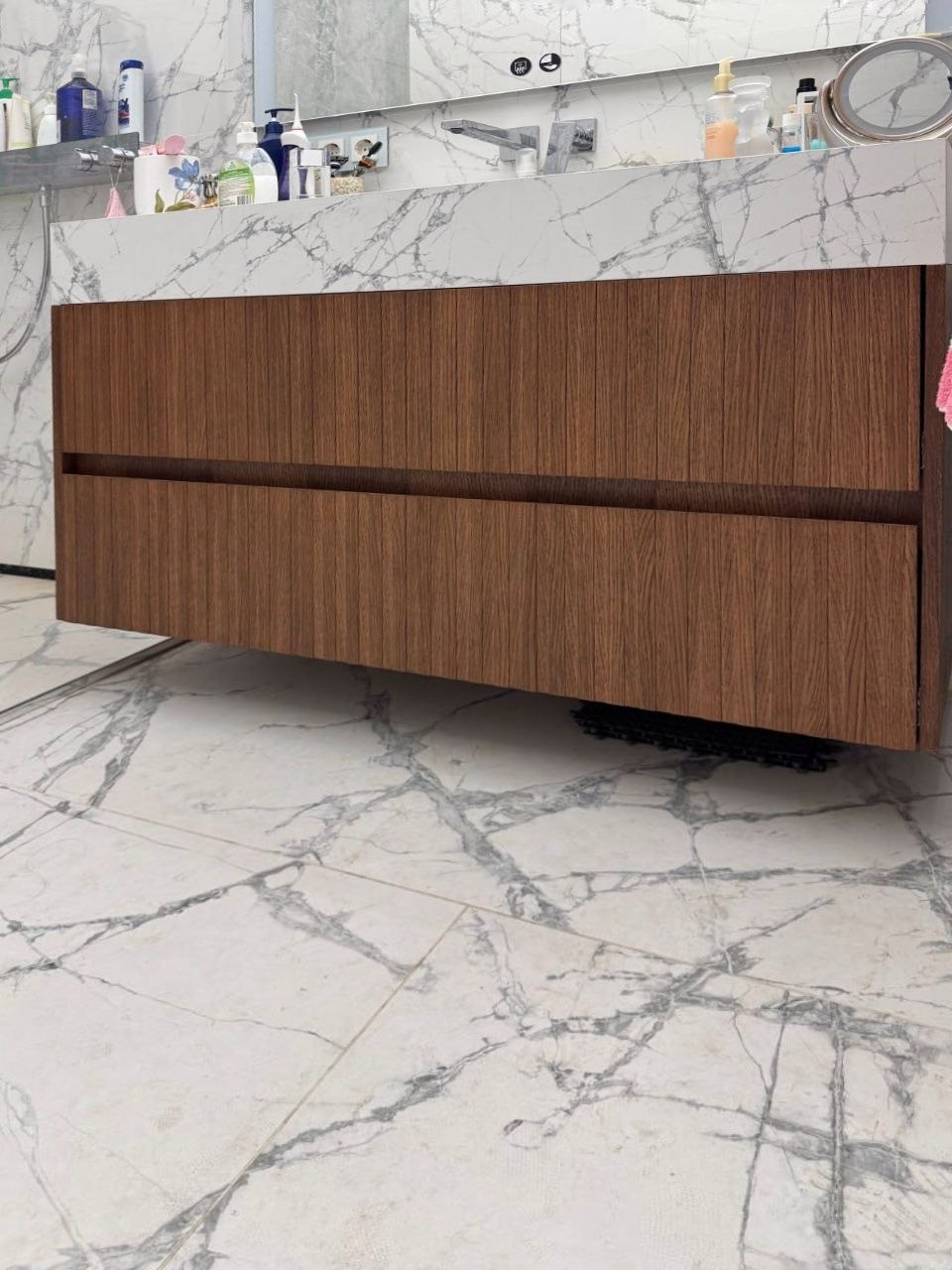 Фрезерованные фасады под шпон — задача, от которой многие мастерские отказываются. Причина проста: сочетание механической обработки и тонкого шпона требует безупречной точности на каждом этапе. Малейшая ошибка — и дорогостоящий материал испорчен. Технологический вызов: Классическая последовательность «сначала фрезеровка, потом шпон» здесь не работает — шпон на рельефной поверхности ложится неравномерно, образуются пузыри и разрывы. Нужно было найти другой путь. Наше решение: Мы разработали обратную технологию. Сначала деталь облицовывается шпоном, и только после этого выполняется фрезеровка. Затем в образовавшиеся пазы вклеиваются полосы палубной доски из дуба. Ювелирная работа: Каждая полоса подбиралась индивидуально, чтобы рисунок текстуры перетекал естественно, без видимых границ и диссонансов. Это требовало художественного взгляда и терпения — иногда на подбор одной вставки уходили часы. Финальный этап — тонирование всей поверхности под образцы существующей мебели в квартире заказчика, чтобы новые фасады органично вписались в интерьер.