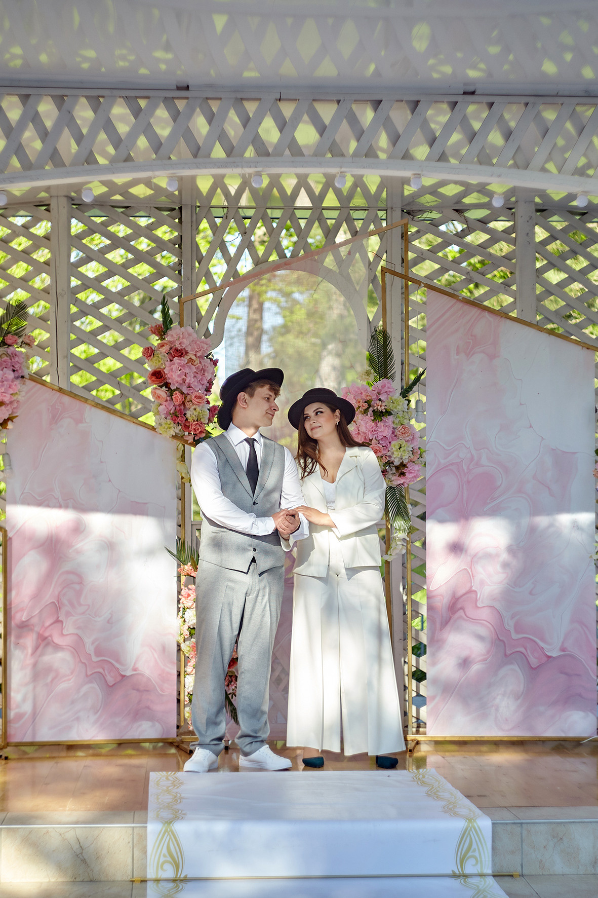 Жених и невеста стоят в летней беседке кафе на церемонии выездной регистрации брака. Стильно. Жених и невеста в шляпах.