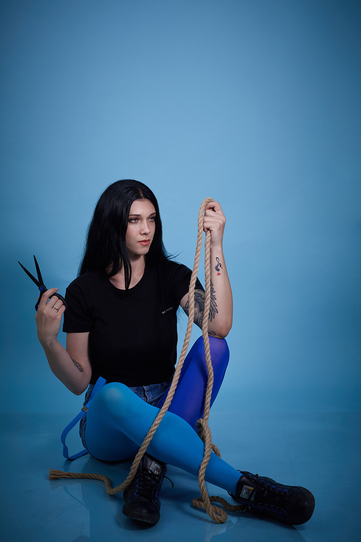 Студийные портрет девушки на голубом фоне. Девушка с татуировками в шортах на подтяжках с веревкой и ножницами в руках. Фотограф. Тимашевск. Кубань. Краснодарский край.