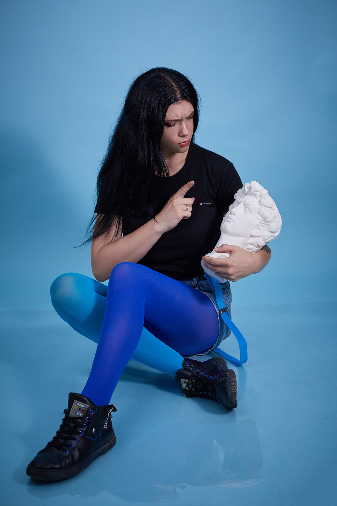 Студийные портрет девушки на голубом фоне. Девушка с татуировками в шортах на подтяжках с гипсовой статуэткой в руках. Фотограф. Тимашевск. Кубань. Краснодарский край.