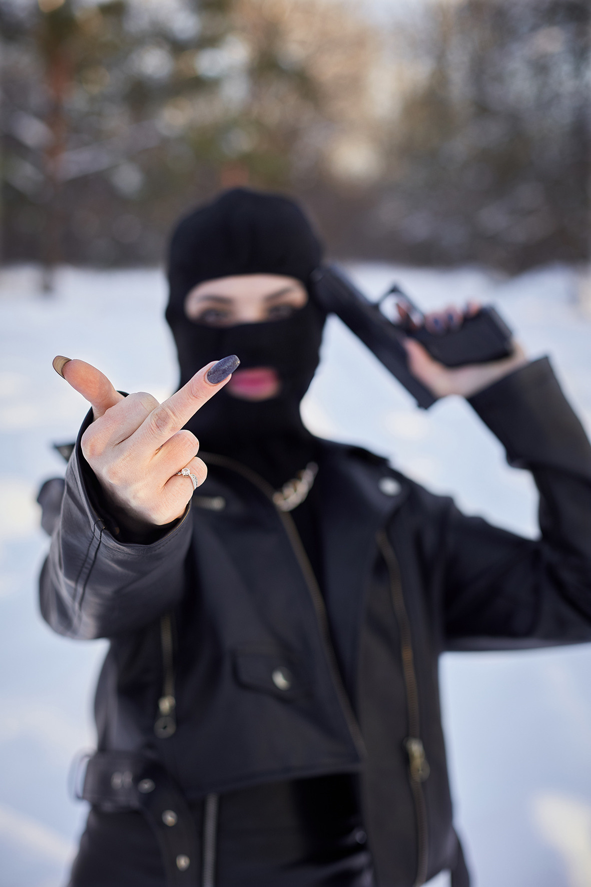 Криминальная дерзкая девушка в кожаной одежде и балаклаве на голове. Девушка с пистолетом показывает неприличный жест. Девушка в образе бандита в заснеженном лесу. Фотограф. Тимашевск. Кубань. Краснодарский край.
