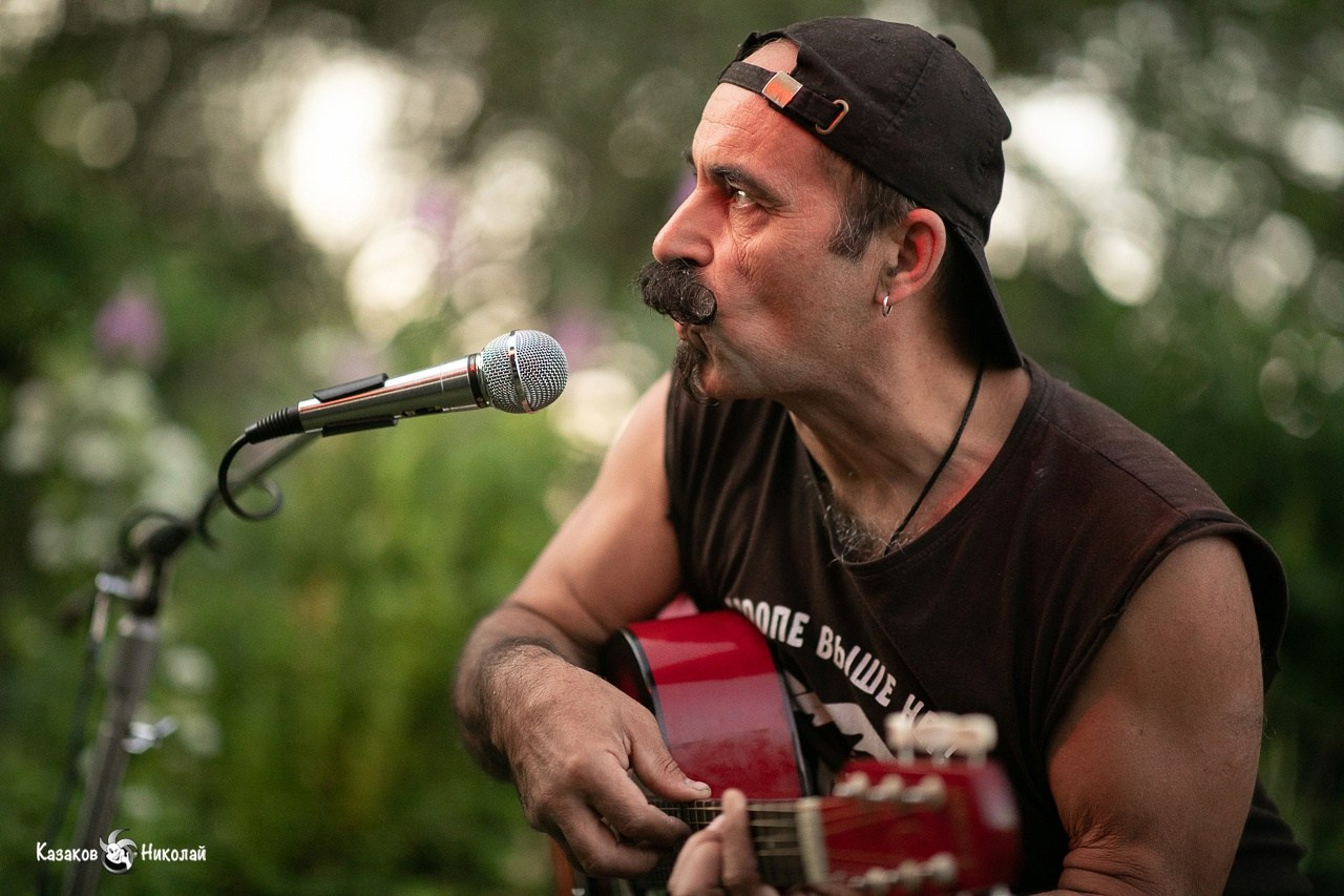Художник и музыкант Михаил Новицкий исполняет авторскую песню под гитару на природе во время квартирного концерта.