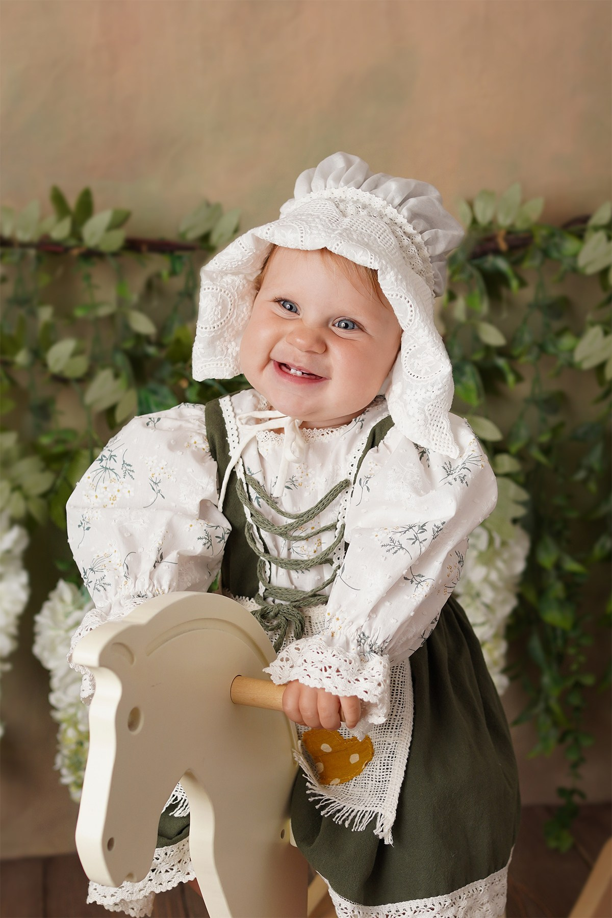 На фото малышка в нарядной одежде и чепчике. Она сидит на игрушечной лошадке.