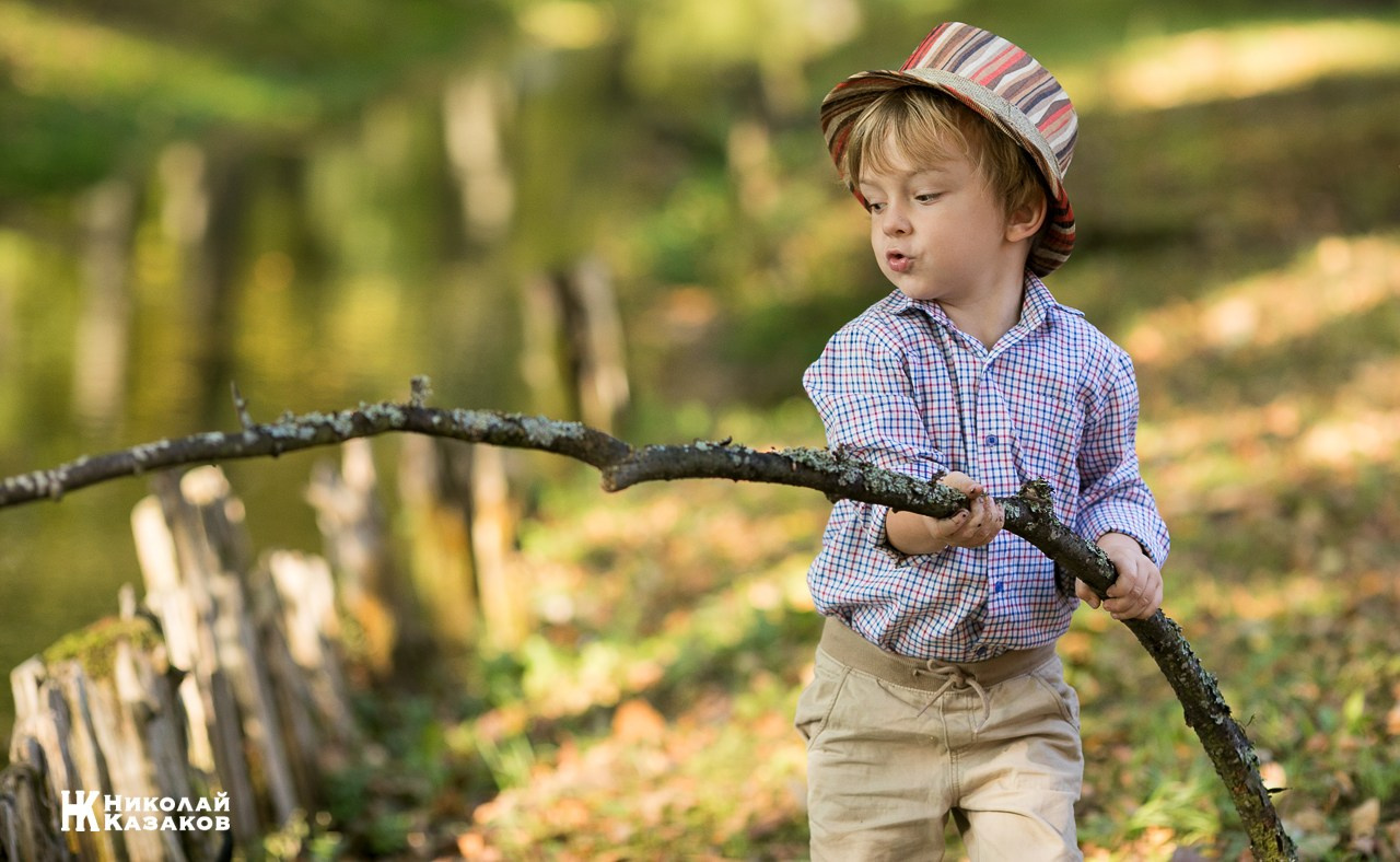Портрет увлеченного маленького мальчика, ловящего палкой водоросли в речке в лучах утреннего солнца