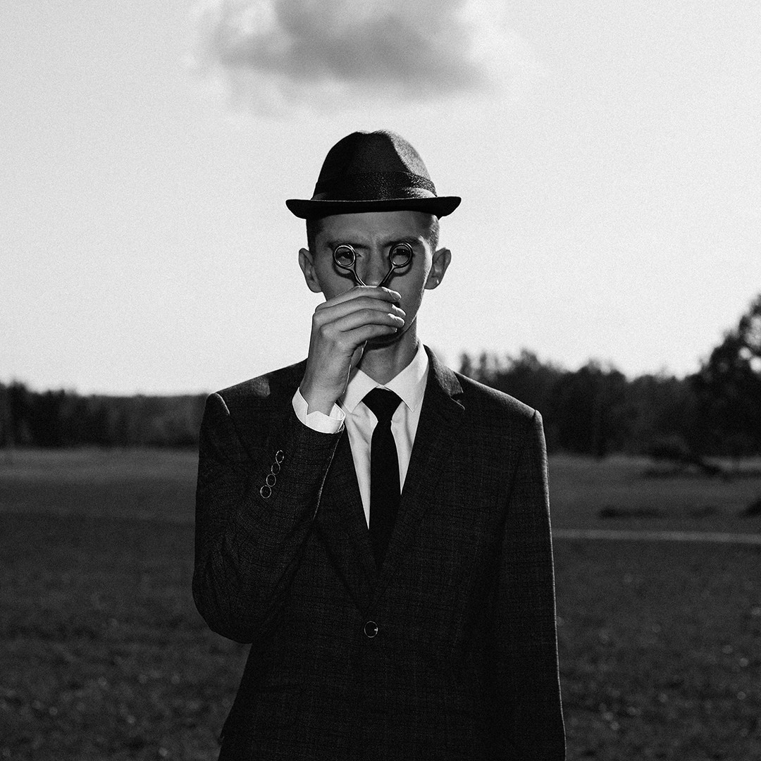 Мужчина в костюме и в шляпе с галстуком позирует в поле в руках у него ножницы которыми он прикрывает лицо