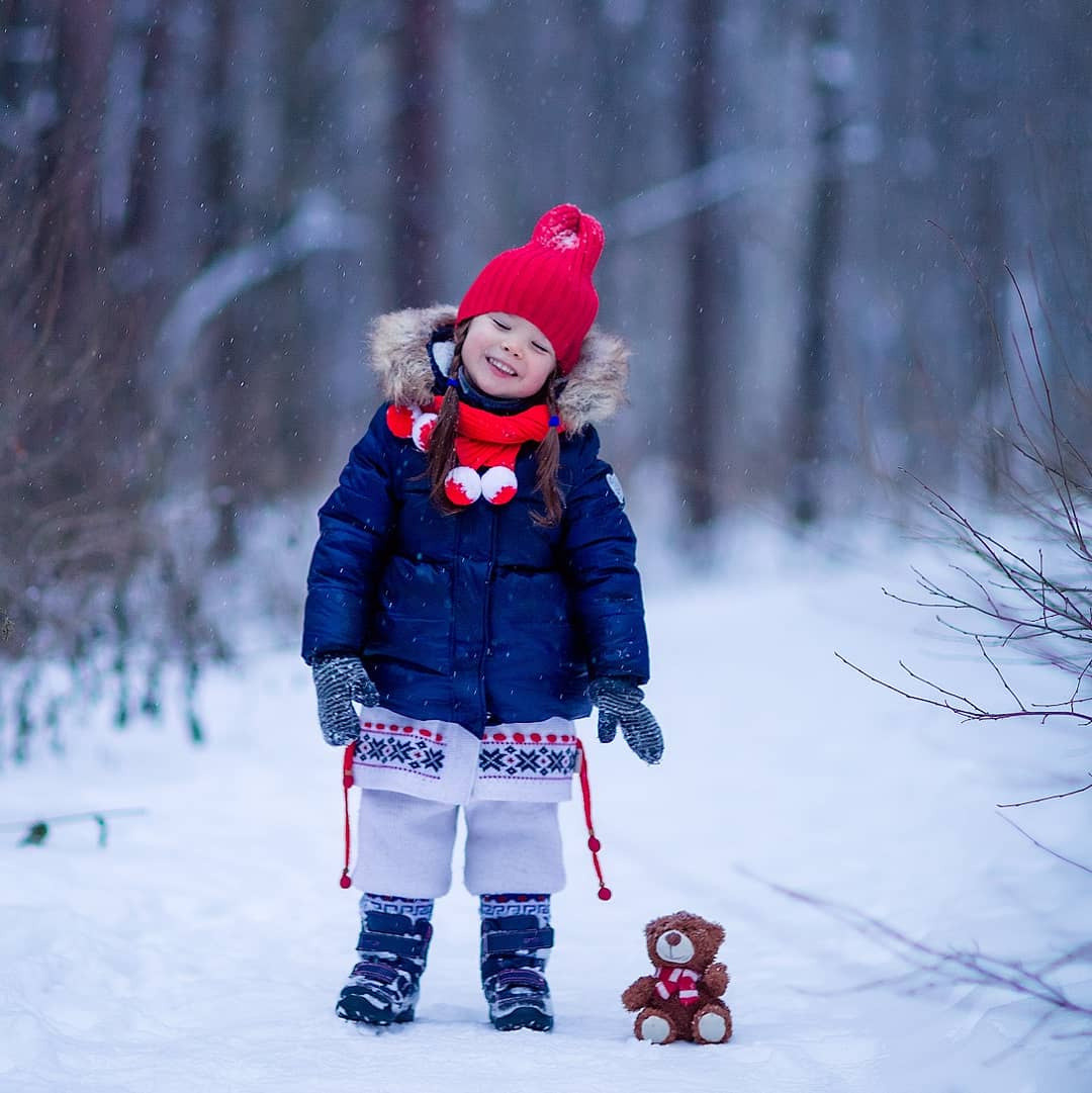 девочка в красной шапке стоит в заснеженном саду с маленькой игрушечной собачкой