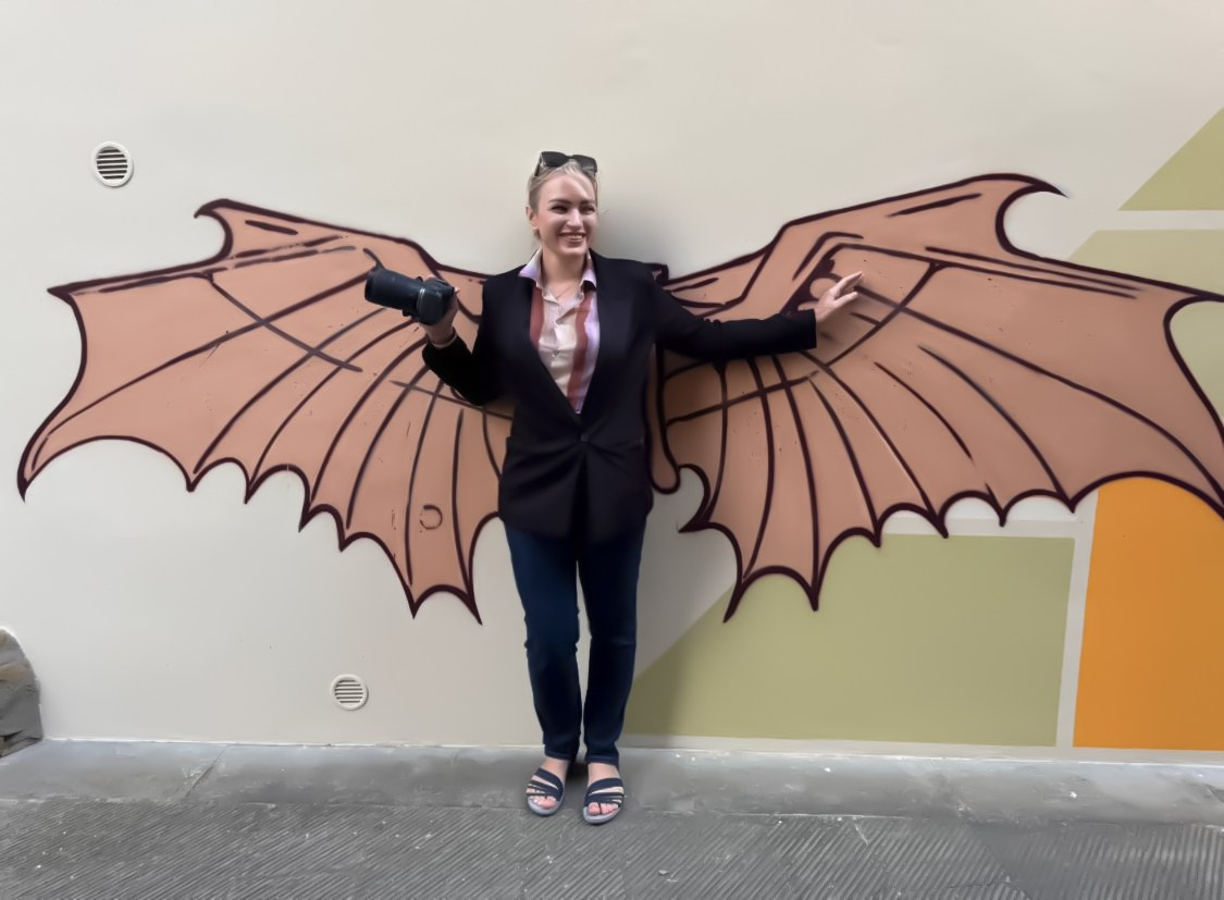 Юлия Перфетти позирует в виде крылатой фигуры на фоне уличной художественной фрески, одетая в блейзер и держащая в руке профессиональную камеру.