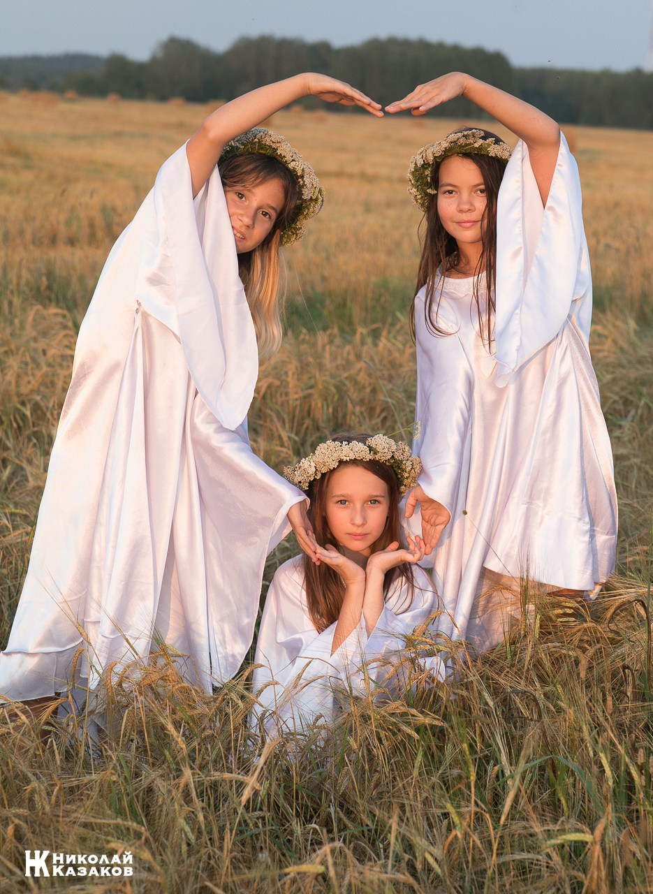 Счастливые девочки подростики в белых платьях создают из рук сердце во время фотосессии в пшеничном поле на закате.