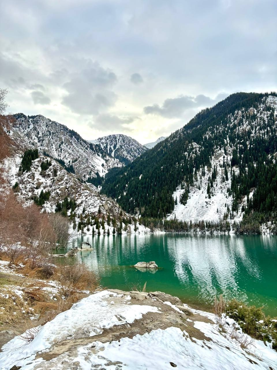 Живописный пейзаж с видом на горное озеро Иссык в Казахстане с изумрудной водой, окруженное снежными горами и еловыми лесами. Небо покрыто облаками, создавая мягкий свет, а в переднем плане виден снег, лежащий на камнях и траве. Атмосфера выглядит свежей и прохладной.