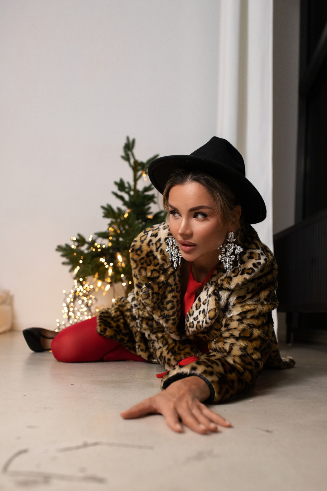 Модная женщина в леопардовой шубе, красном наряде и черной шляпе позирует на полу у новогодней елки с гирляндами в стильном интерьере.