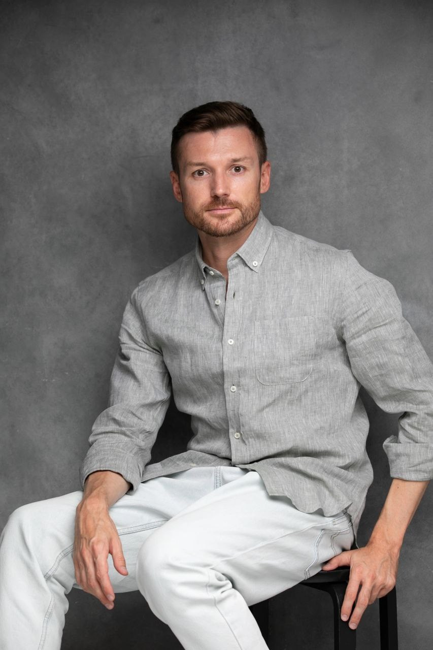 Мужчина с короткой щетиной в серой рубашке и светлых джинсах на фоне серой стены