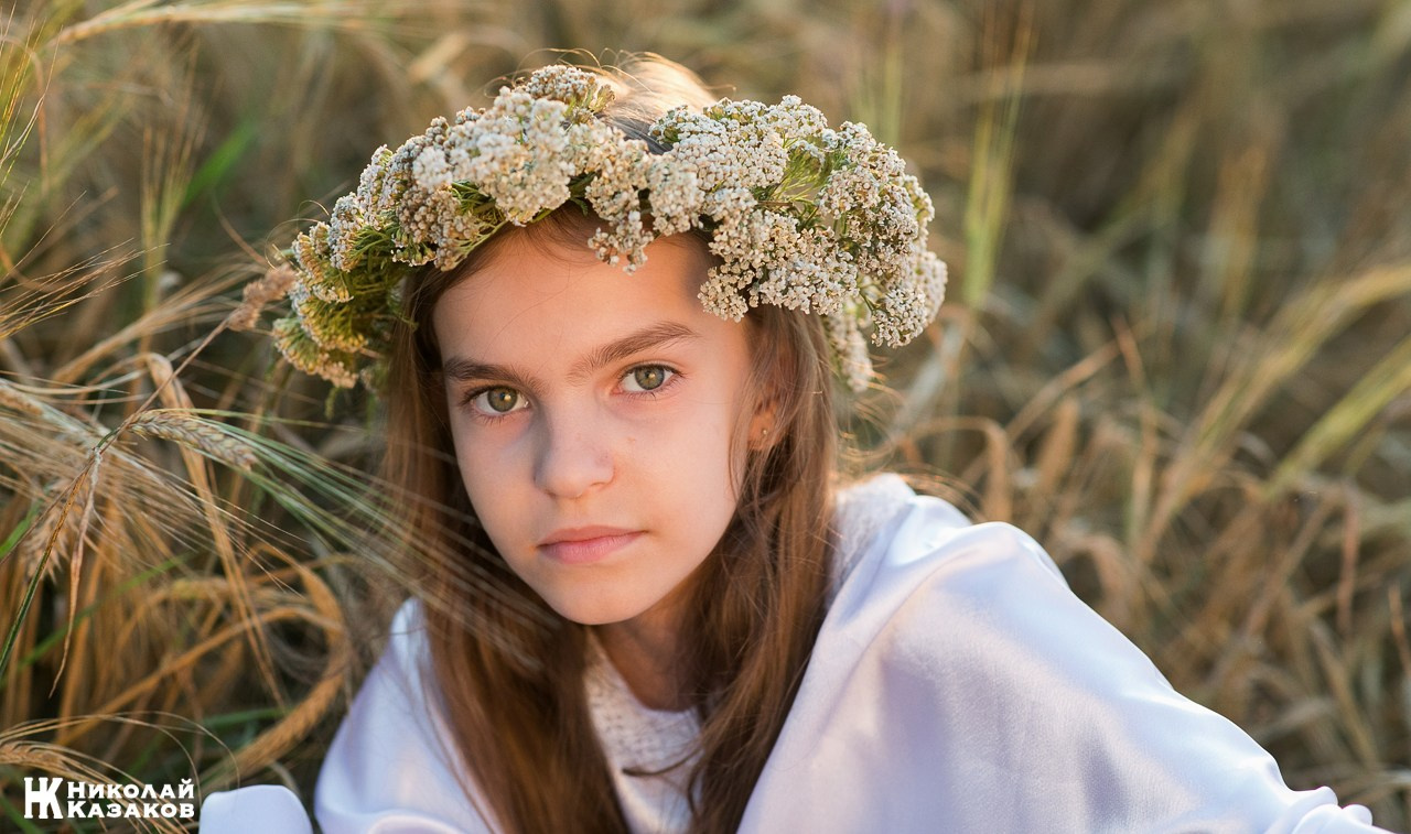 Крупный портрет нежной девочки с венком на голове в лучах заката, лежащая в поел пшеницы во время летней фотосессии.