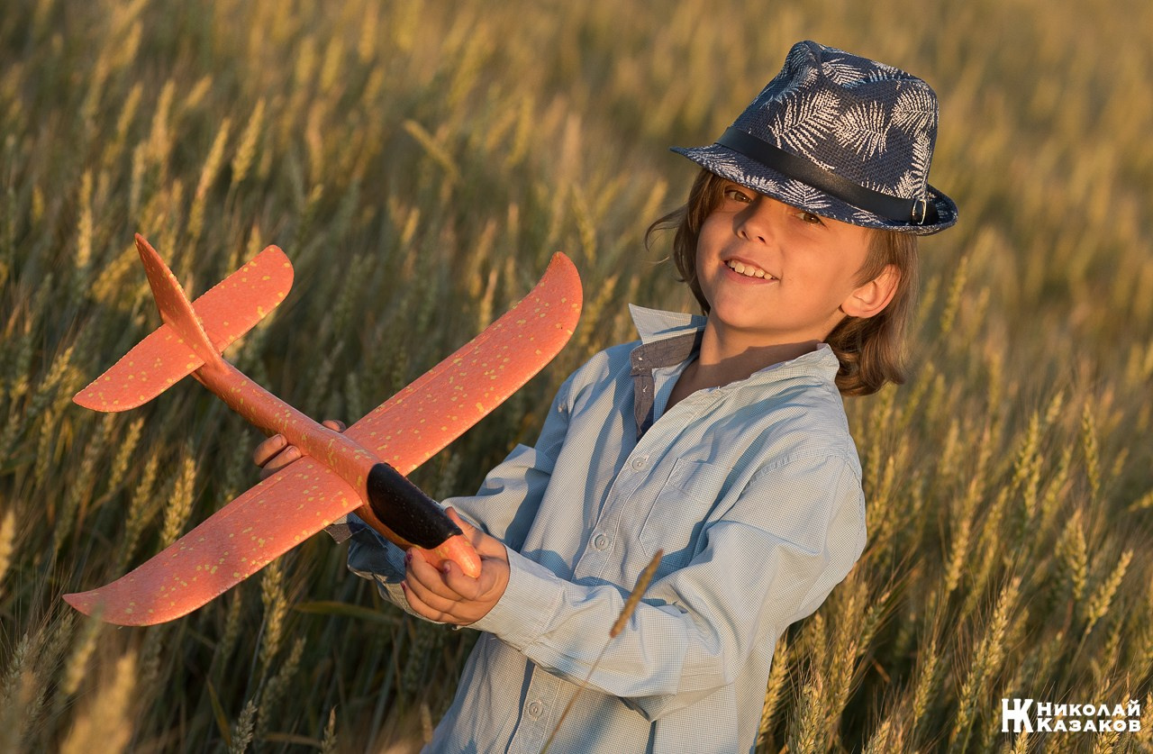 Счастливый маленький мальчик с светлыми волосами идет по пшеничному полу в лучах закатного солнца , держа в руках игрушечный самолет.