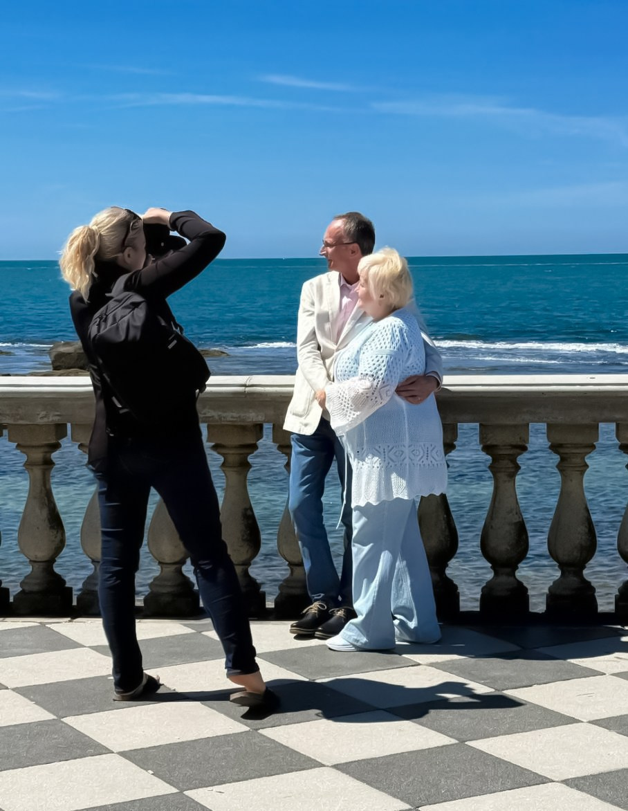 Фотограф Юлия Перфетти в черном делает портрет пары на клетчатой набережной на фоне синего моря и неба.