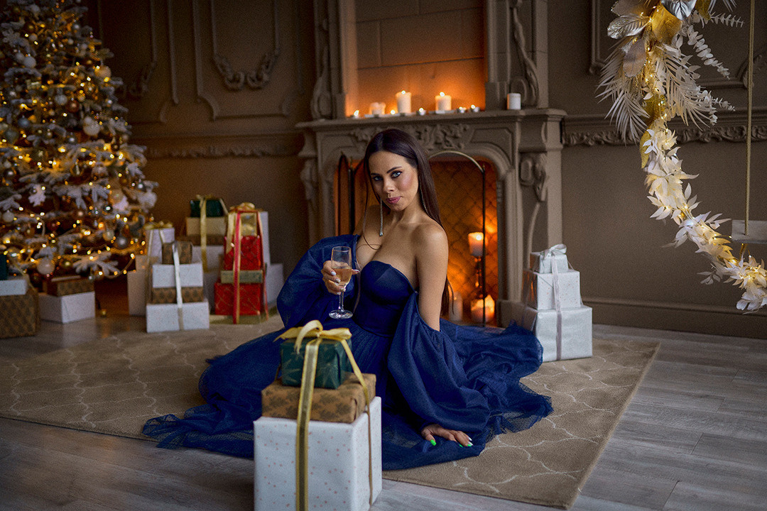 Девушка в вечернем платье сидит в комнате с бокалом вина возле камина, комната украшена к новогоднему празднику