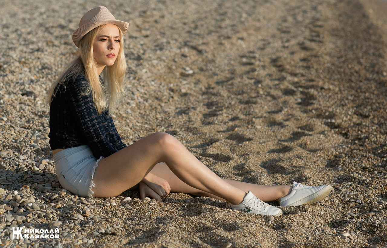 Стильная молодая женщина сидит на пляже в лучах солнца и смотрит на волны.