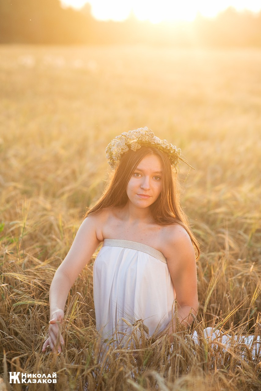 Красивая девушка в лучах закатного солнца , сидящая в пшеничном поле во время фотосессии.