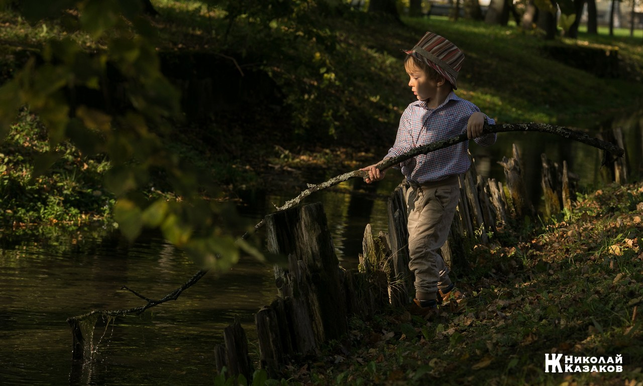 Портрет увлеченного маленького мальчика, ловящего палкой водоросли в речке в лучах утреннего солнца