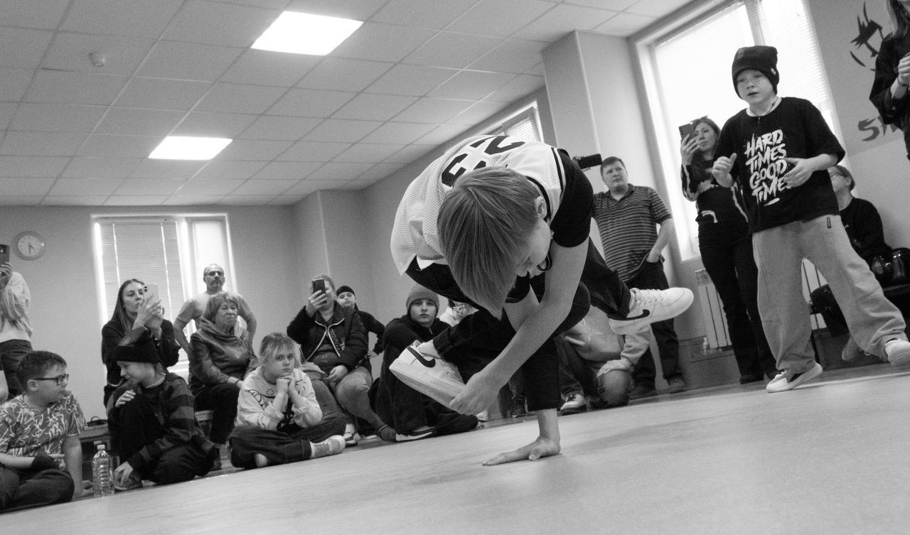 Чёрно-белое фото: молодой брейкер выполняет freeze на полу танцевального зала, окружённый зрителями разных возрастов; некоторые фотографируют на телефоны; документальная съёмка уличной культуры и брейк-данса