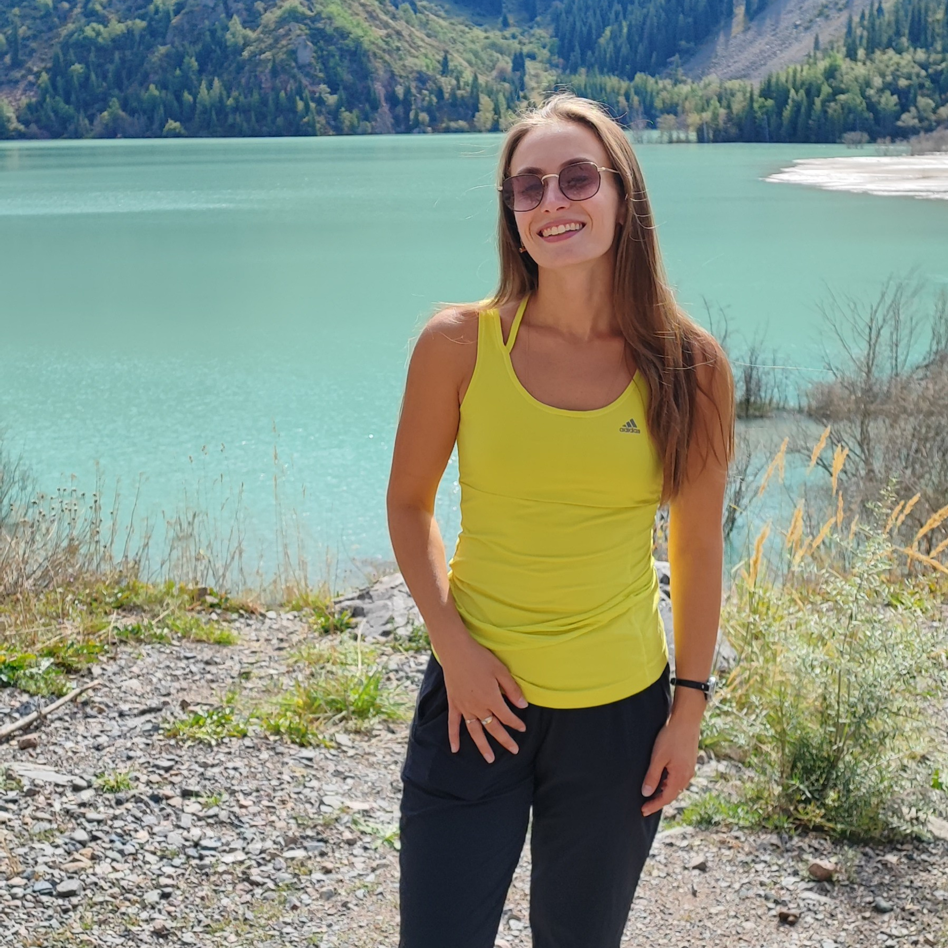 На фото улыбающаяся девушка в жёлтой спортивной майке и чёрных брюках стоит на фоне бирюзового горного озера Иссык, окружённого зелёными склонами с густыми хвойными лесами. Она носит солнцезащитные очки и фиолетовые кроссовки, создавая контраст с природным пейзажем. Светлое небо и солнечные лучи подчёркивают яркость воды и естественную красоту гор.