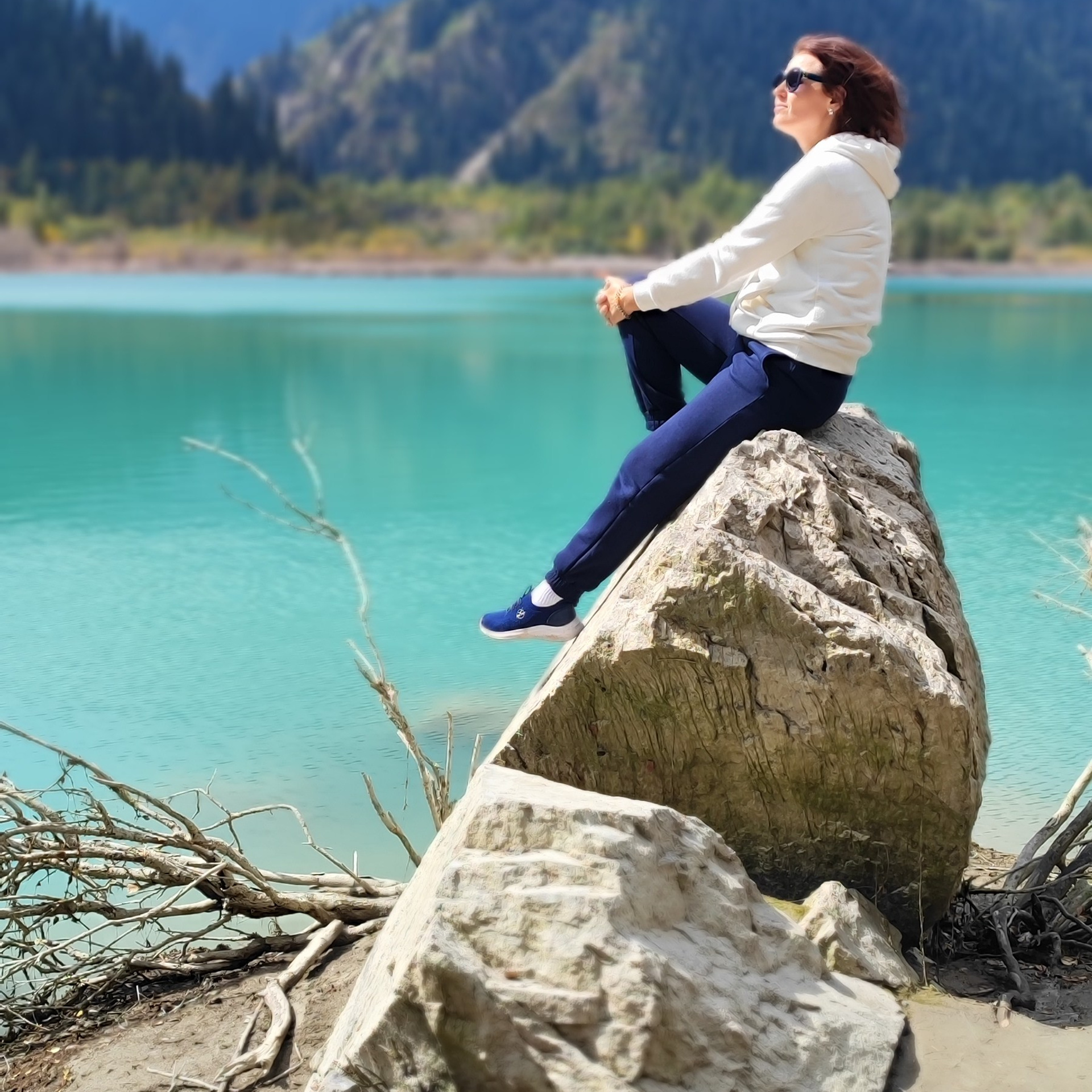 Женщина в белой толстовке и синих брюках сидит на большом камне у бирюзового озера Иссык в Алматы. Она смотрит вдаль, наслаждаясь природой и свежим воздухом. За её спиной видны величественные горы, покрытые зеленью, под серо-голубым небом с облаками. Атмосфера спокойствия и умиротворения.