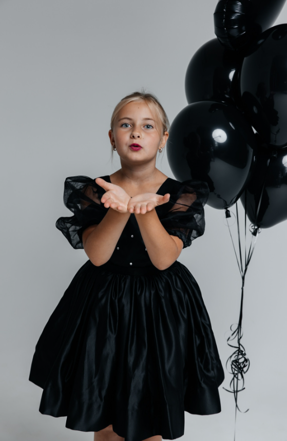 Девочка в чёрном платье позирует с жестом «поцелуй», рядом — чёрные шары. Фон нейтральный.