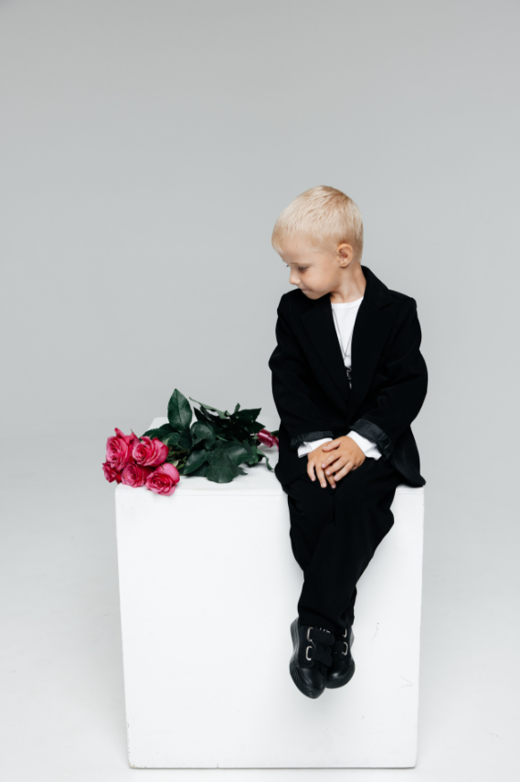 Малыш в чёрном костюме сидит на белом кубе, рядом — букет розовых роз.