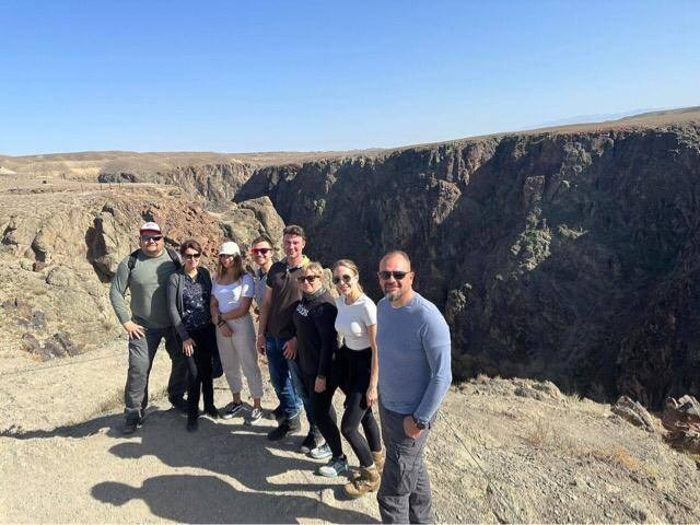 Группа из восьми человек позирует на фоне глубокого Черного каньона в районе Алматы в Казахстане под ясным голубым небом. Все участники одеты в удобную повседневную одежду для прогулки на природе. Скалы Черного каньона впечатляюще подчеркивают масштаб ландшафта, а сухие холмы на горизонте создают контраст с каменистыми обрывами. Светлое утро и яркое солнце добавляют фотографии теплую атмосферу.