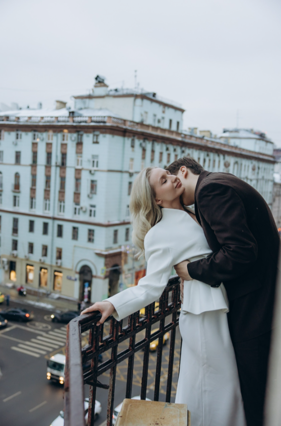 Пара на балконе: парень в тёмном костюме обнимает девушку в белом платье, целует её в щёку. За ними — городской пейзаж.