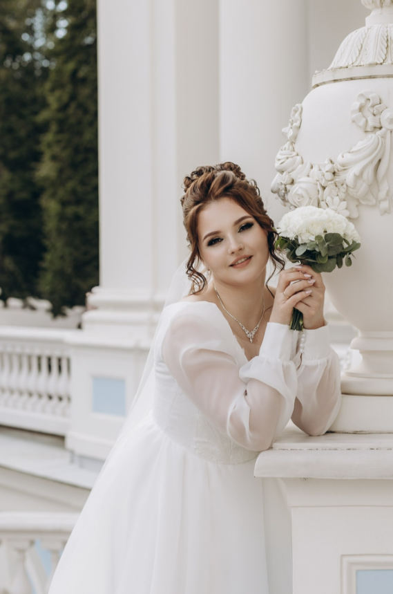 Невеста в белом платье с букетом нежно опирается на колонну. Место похоже на усадьбу.