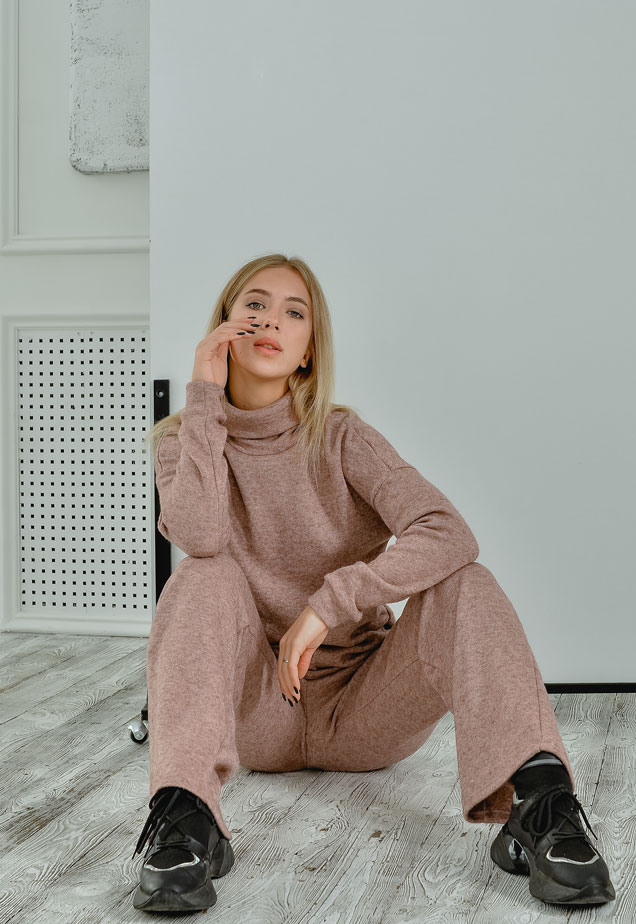 Девушка в свитере и штанах сидит на полу в светлой фотостудии.