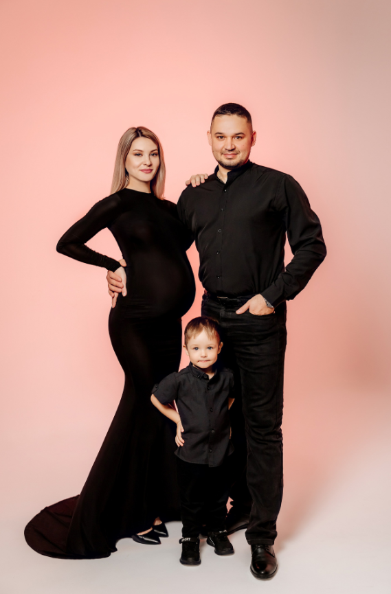 Беременная женщина в длинном чёрном платье, мужчина в чёрной рубашке и маленький ребёнок. Фон нежно-розовый, атмосфера стильная и тёплая.