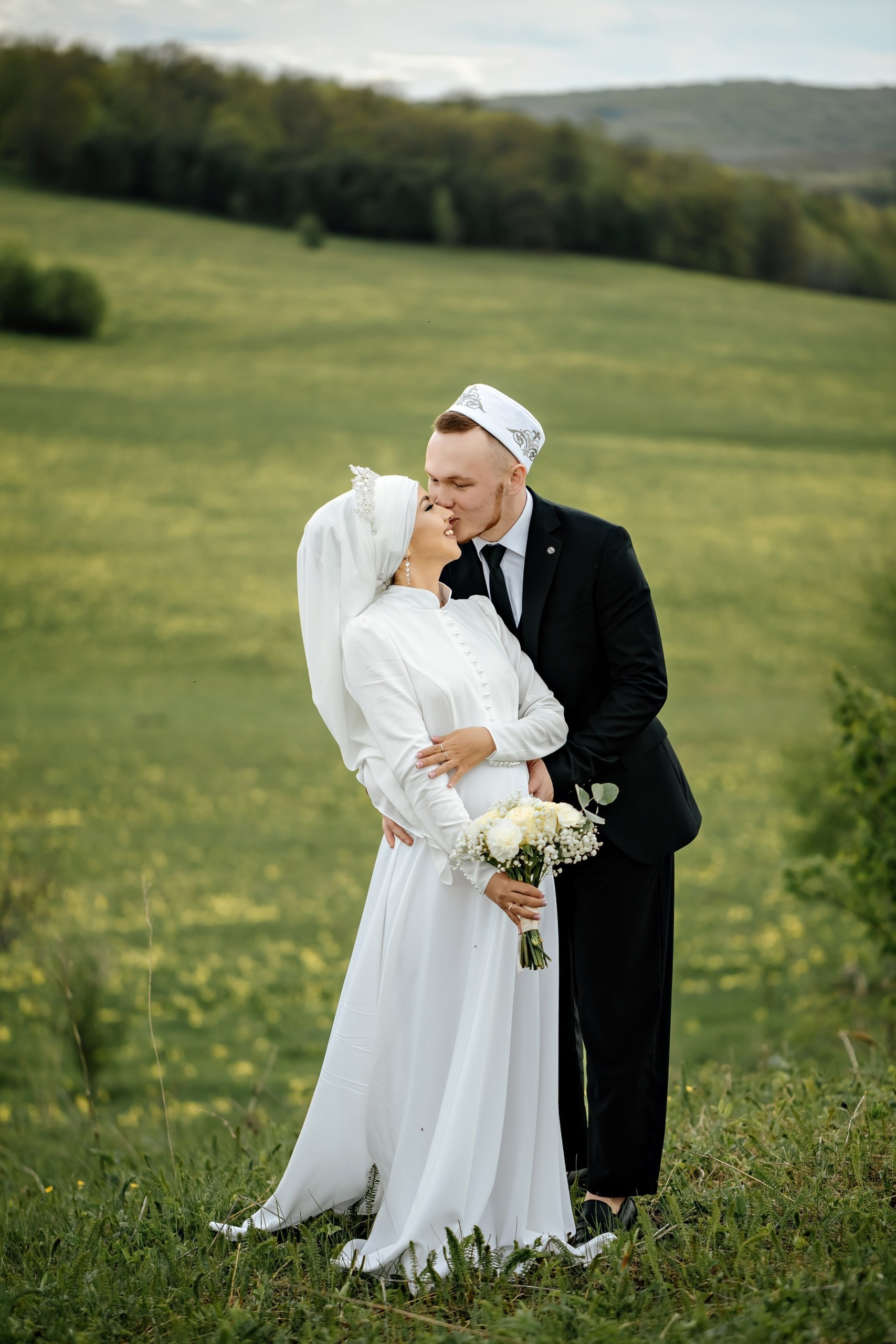 Никах. Пара в свадебных нарядах — жених в чёрном костюме и тюбетейке, невеста в белом платье с покрывалом. Стоят на зелёном лугу, обнимаются, атмосфера нежная и романтичная.