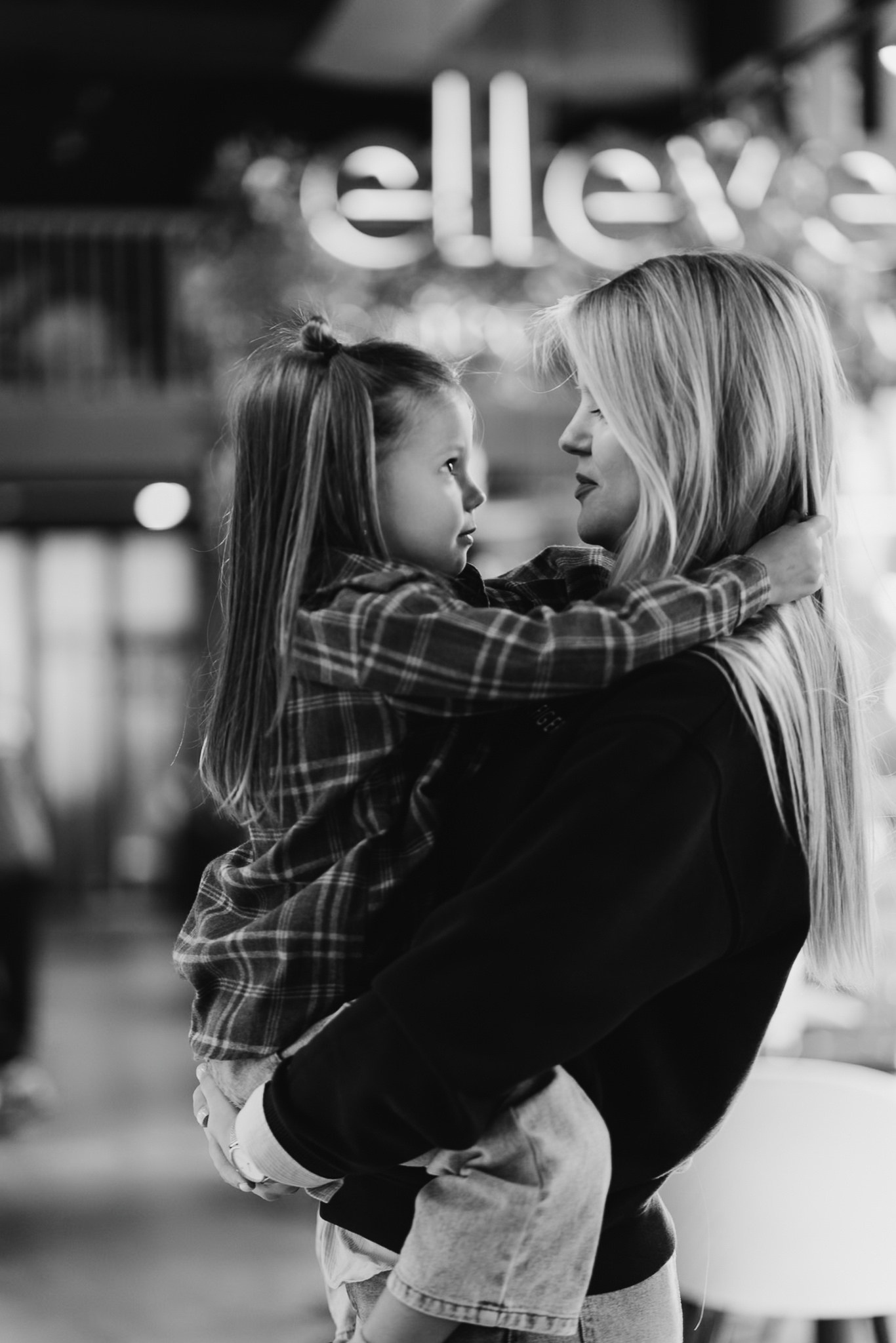 На чёрно-белом фото мама держит на руках дочку, они смотрят друг на друга. На фоне — светящаяся вывеска «ellev»