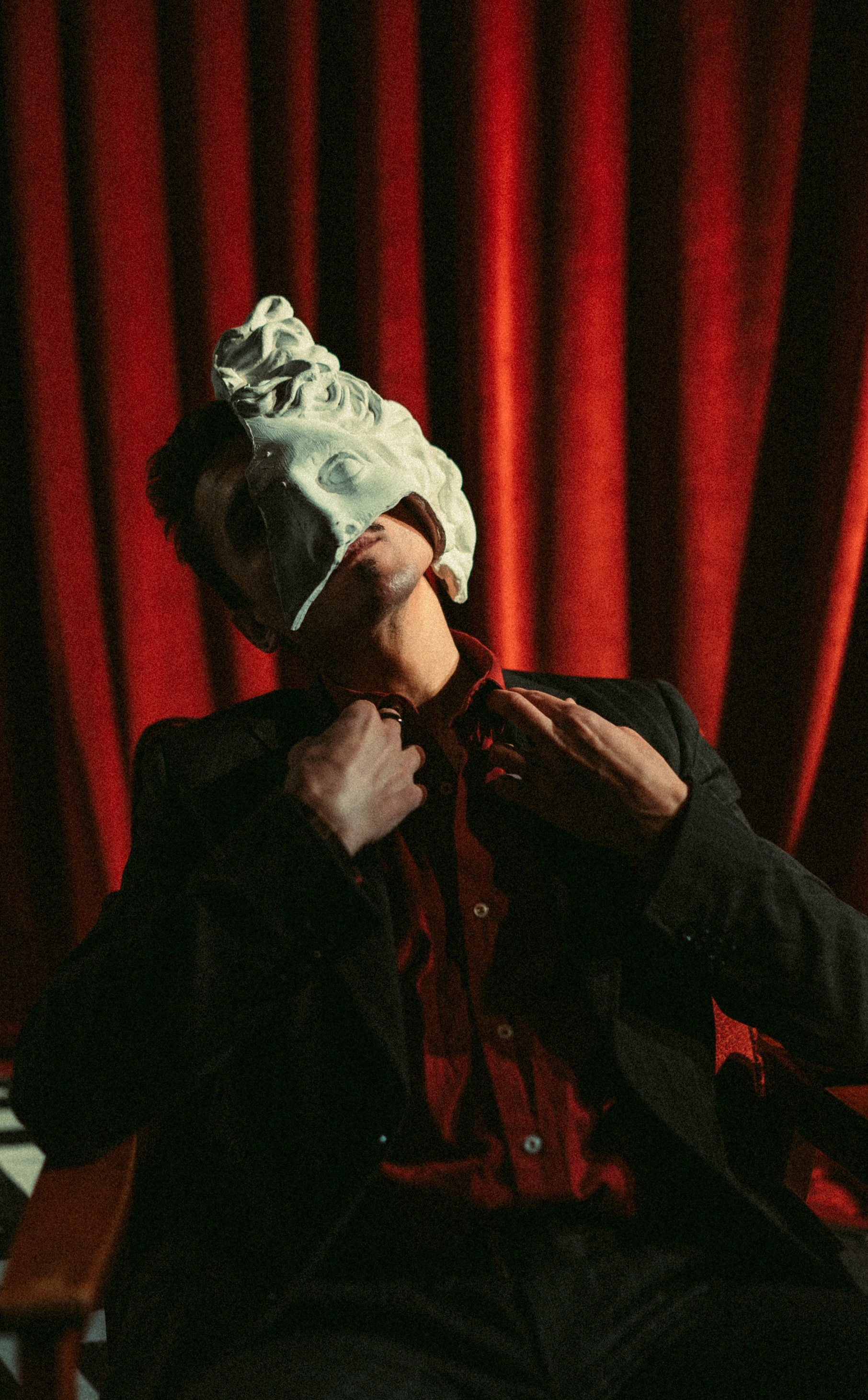 Мужчина в тёмном костюме с гипсовой маской на лице сидит перед красным бархатным занавесом в театральной атмосфере
