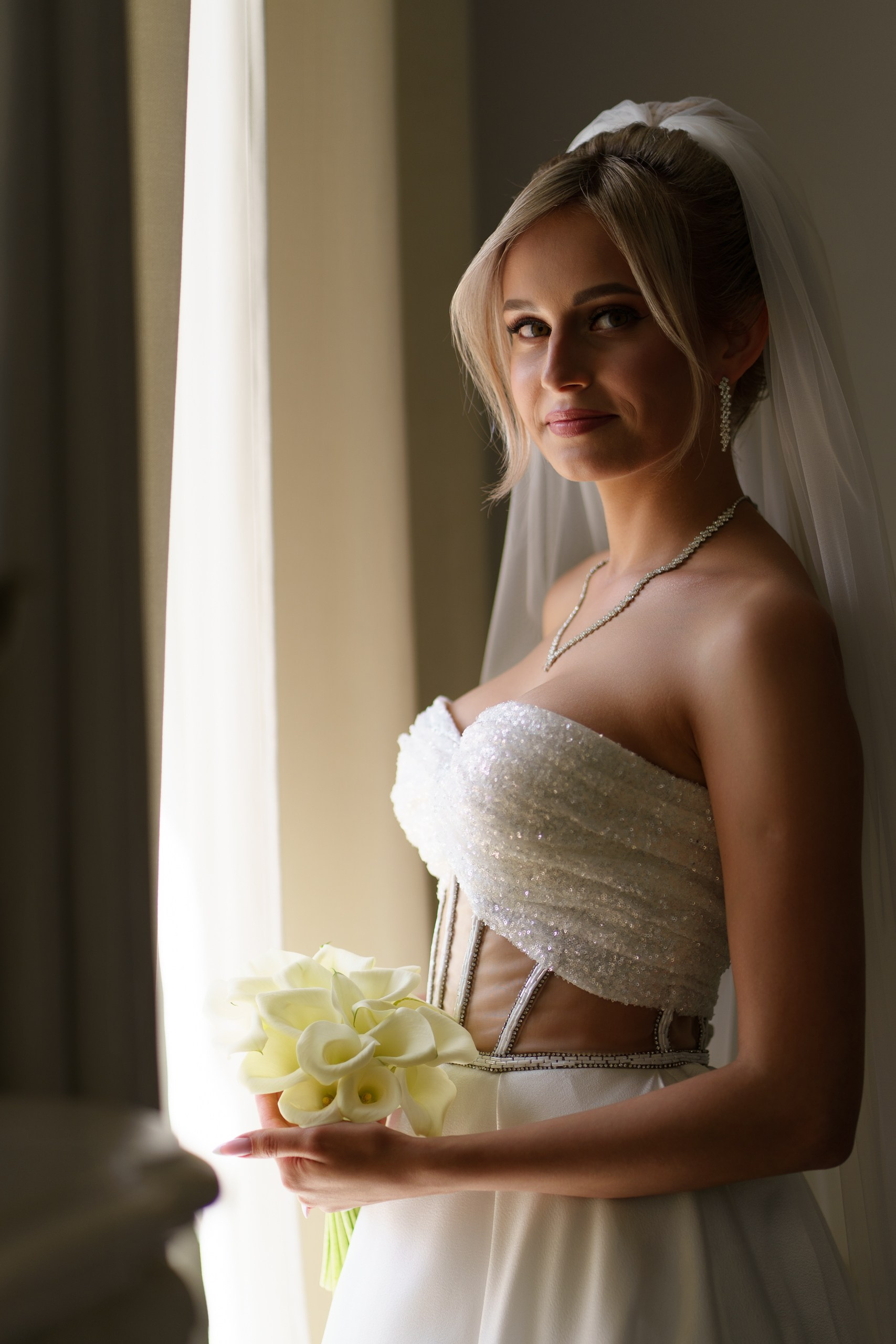 Молодая невеста в сверкающем свадебном платье позирует у окна. Она держит букет нежных калл, её волосы аккуратно уложены, а фата изящно ниспадает за спиной | свадебная фотография Екатеринбург