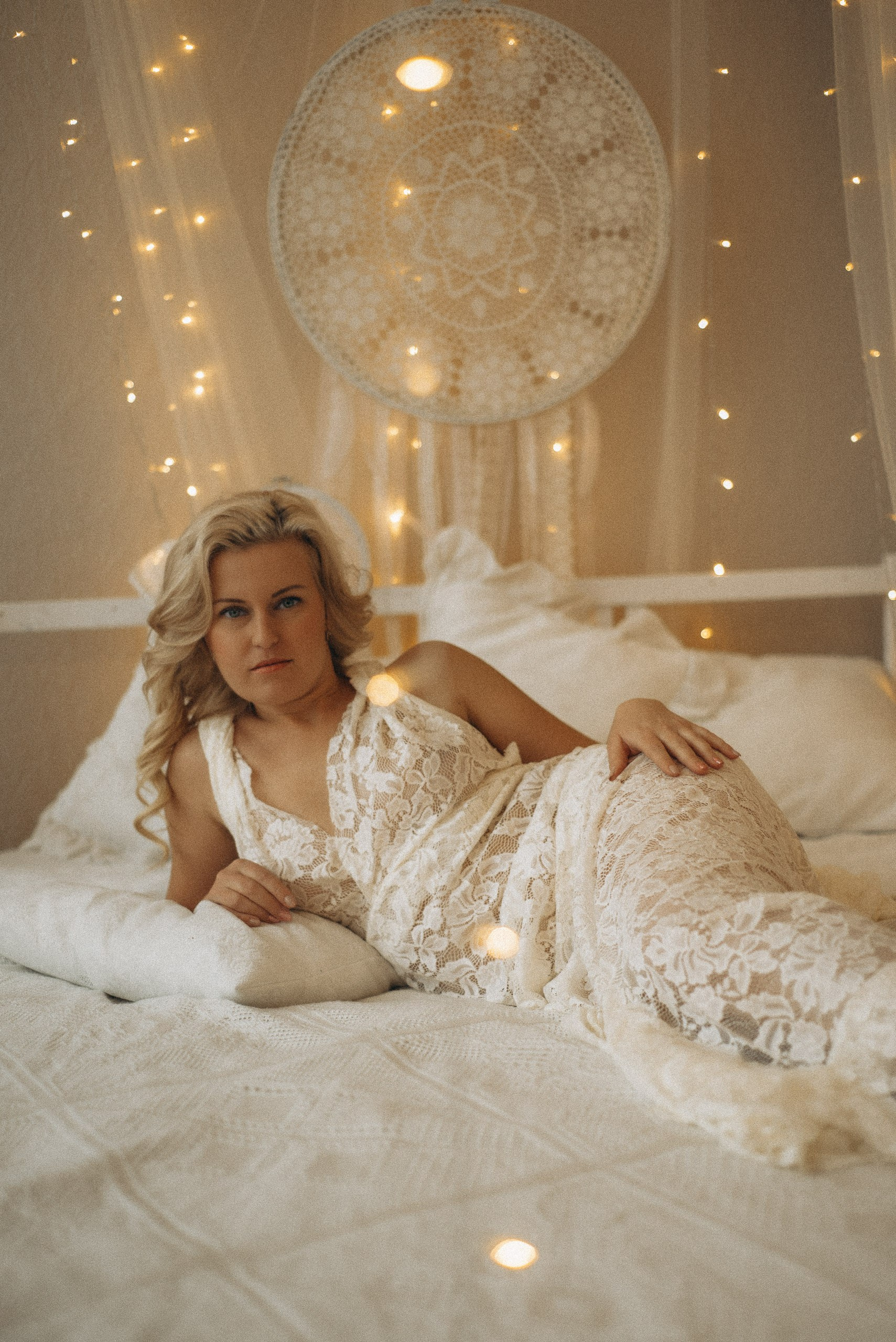 Женский портрет на кровати в кружевном платье. Натали Каролин смотрит в кадр, мягкое освещение от огоньков на переднем плане. Эстетика будуарной фотосессии в стиле бохо
