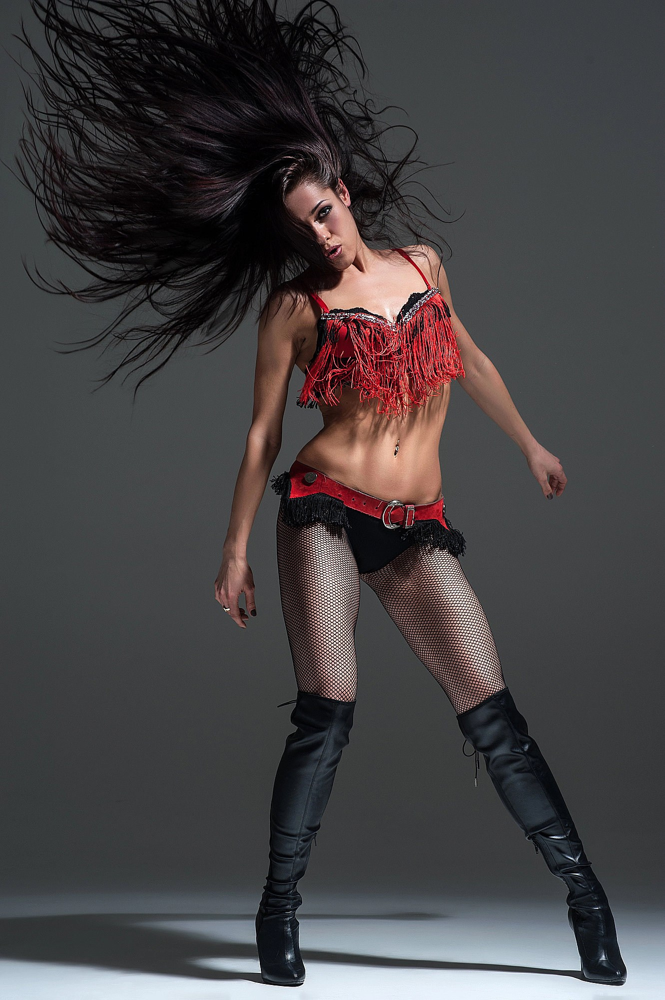 Танцовщица в красном топе резко выбрасывает волосы назад, создавая эффект взрыва энергии. Чёрные ботфорты и уверенная стойка добавляют характер сценического шоу. Студийный художественный портрет, Москва и Истринский район.