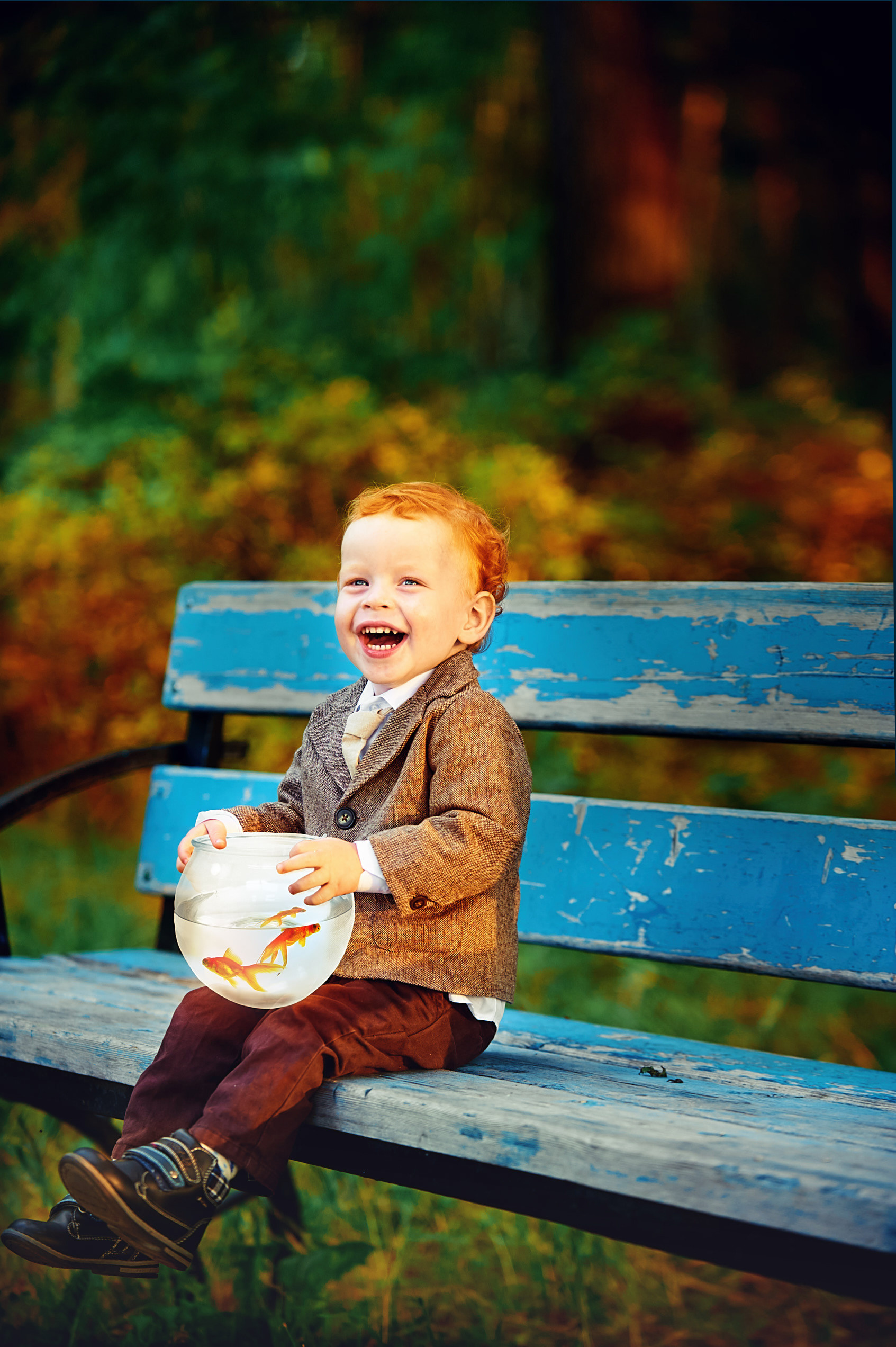 рыжеволосый малыш сидит на синей лавочке с аквариумом с одной рыбкой и смеется